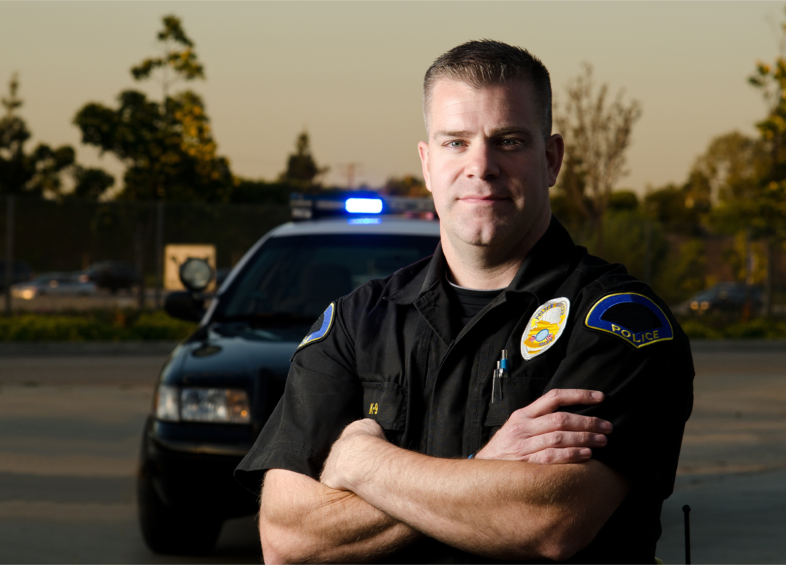 Male police officer with his arms crossed standing in front of his police car